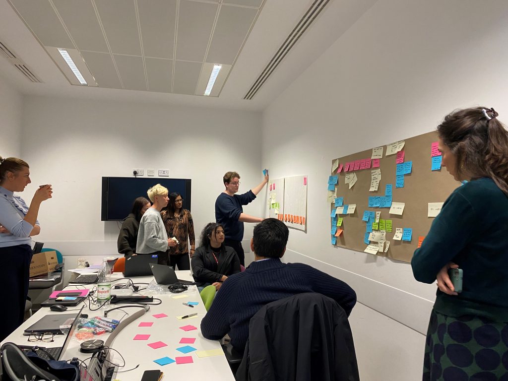 This is an image showing a diverse group of individuals during a workshop. They are discussing the customer journey, placing coloured post it notes onto a wall to map out the step by step journey and issues and pain points from a service area perspective.