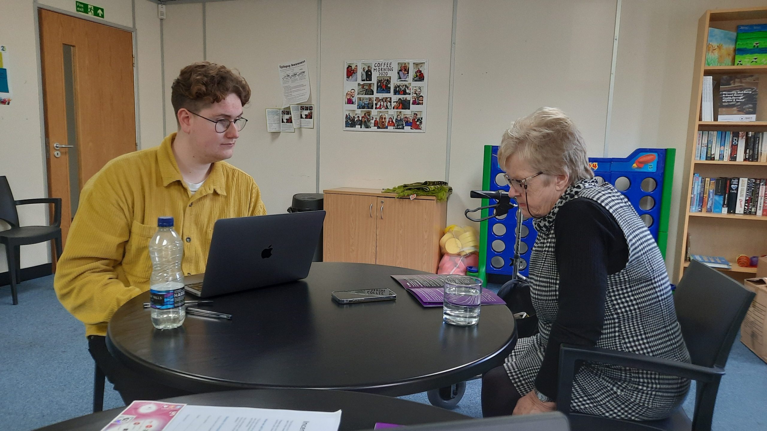 This is an image of two people, one young male and one older lady sitting at a table. The older lady is reading a leaflet.