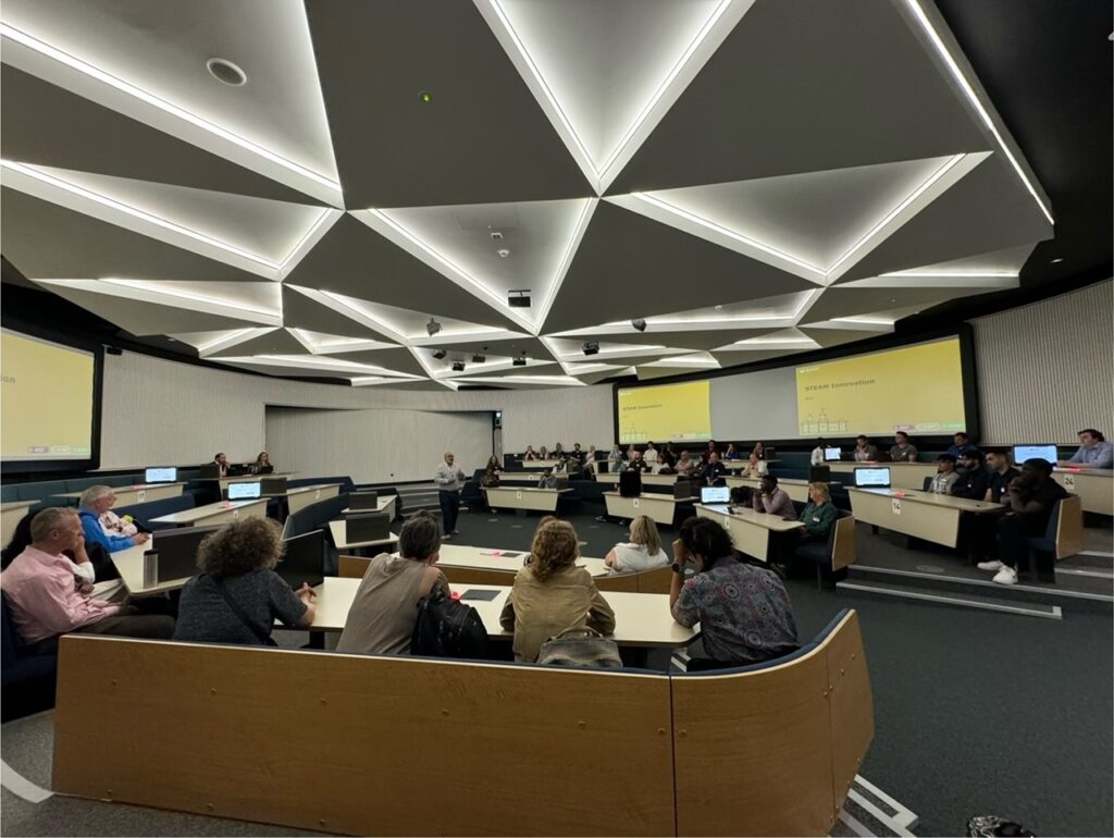 This image shows Professor Rehan Bhana talking to an audience of people seated in an auditorium about the STEAMHouse initiative