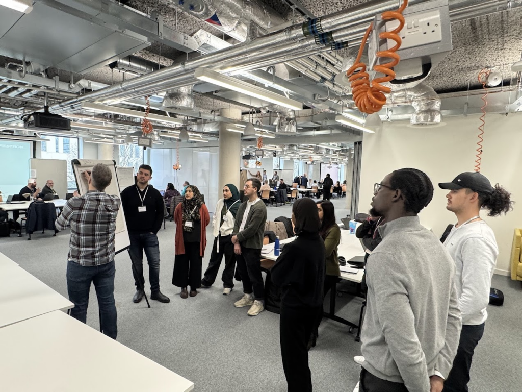 This is an image showing a group of people of mixed genders, age groups and cultures standing in a group discussing their hack problem